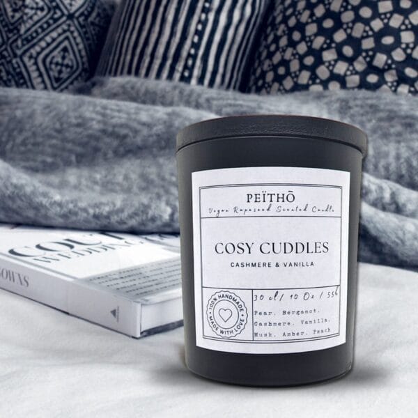 Peitho-Perfumes.ScentedCandles_ A candle with the words cozy cuddles sitting on top of a bed.